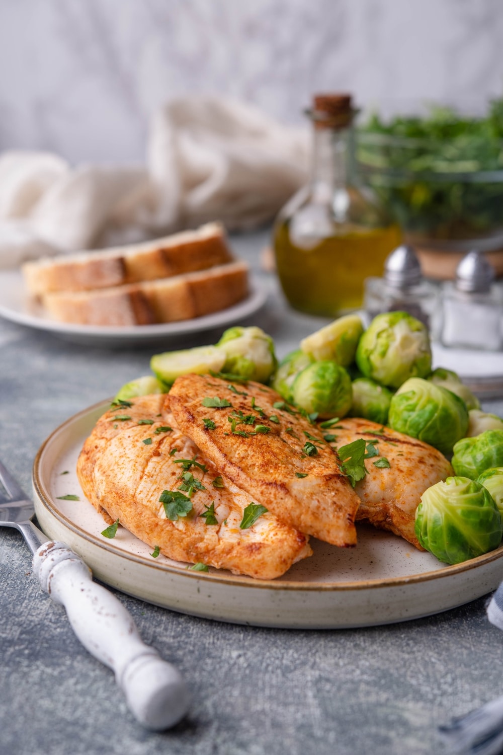 Sauteed chicken breasts on a plate, garnished with herbs and served with steamed brussel sprouts. Behind is a plate of sliced bread, a bottle of olive oil, a glass bowl filled with greens, and some salt and pepper shakers.