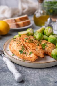 Sauteed chicken breasts garnished with herbs on a plate with steamed brussel sprouts. A fork with a white handle is next to the plate. In the background is a lemon, a plate of sliced bread, a bottle of olive oil, salt and pepper shakers, and a glass bowl of greens.