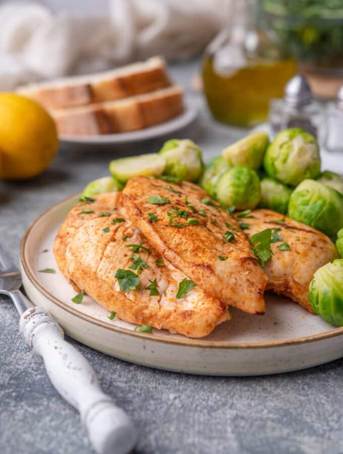 Sauteed chicken breasts garnished with herbs on a plate with steamed brussel sprouts. A fork with a white handle is next to the plate. In the background is a lemon, a plate of sliced bread, a bottle of olive oil, salt and pepper shakers, and a glass bowl of greens.