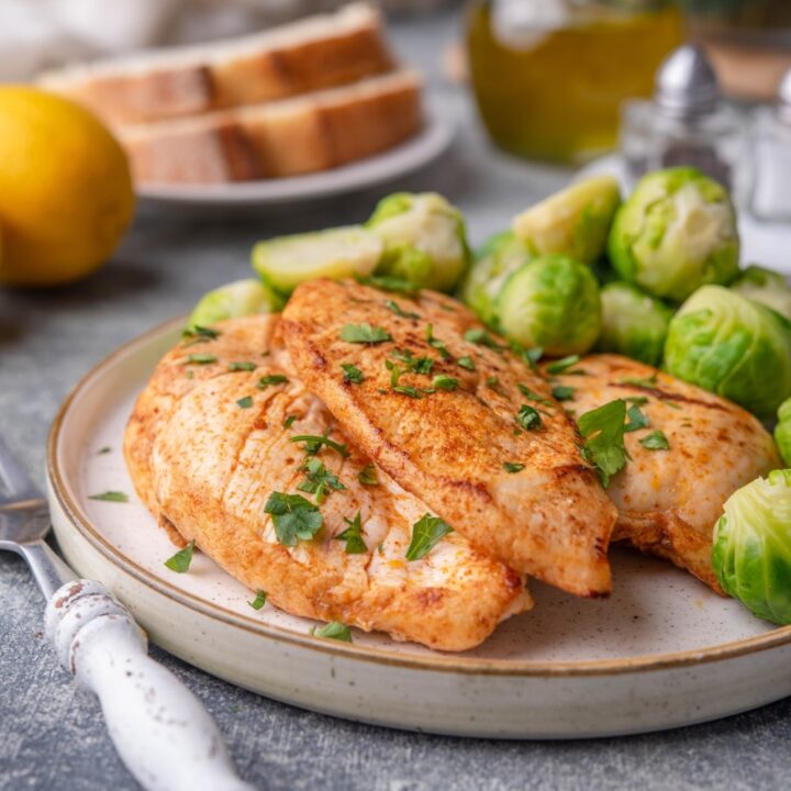 Sauteed chicken breasts garnished with herbs on a plate with steamed brussel sprouts. A fork with a white handle is next to the plate. In the background is a lemon, a plate of sliced bread, a bottle of olive oil, salt and pepper shakers, and a glass bowl of greens.