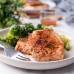 Sauteed pork chops served with steamed broccoli on a white plate, next to a mini pitcher of pan sauce. Another plate of sauteed pork chops with broccoli is in the back.