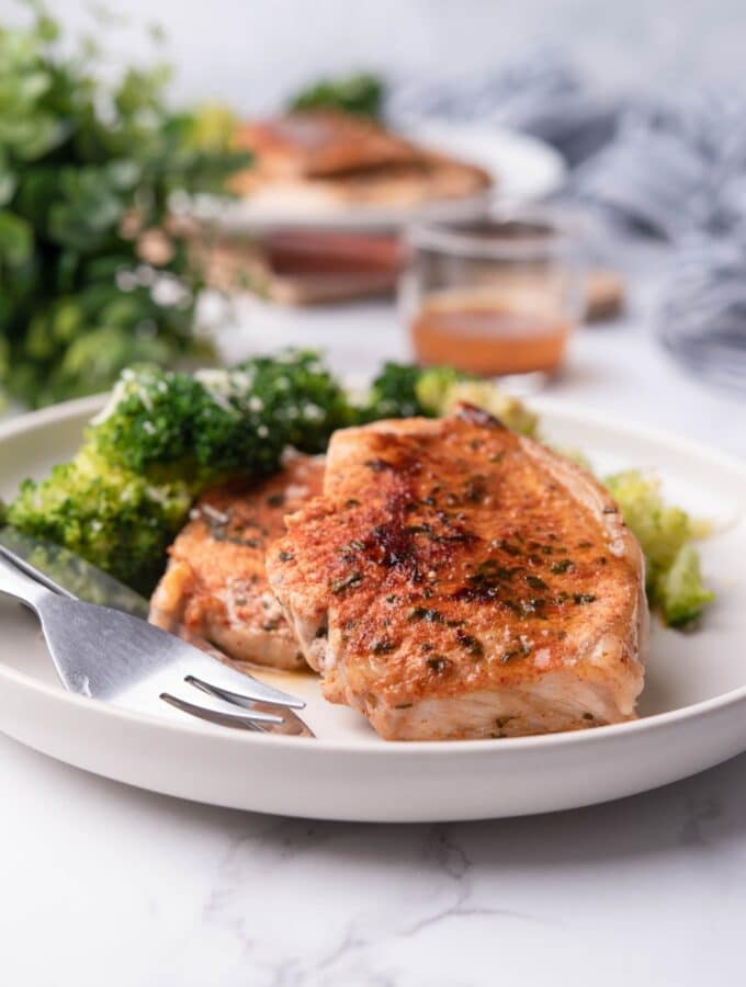 Sauteed pork chops served with steamed broccoli on a white plate, next to a mini pitcher of pan sauce. Another plate of sauteed pork chops with broccoli is in the back.