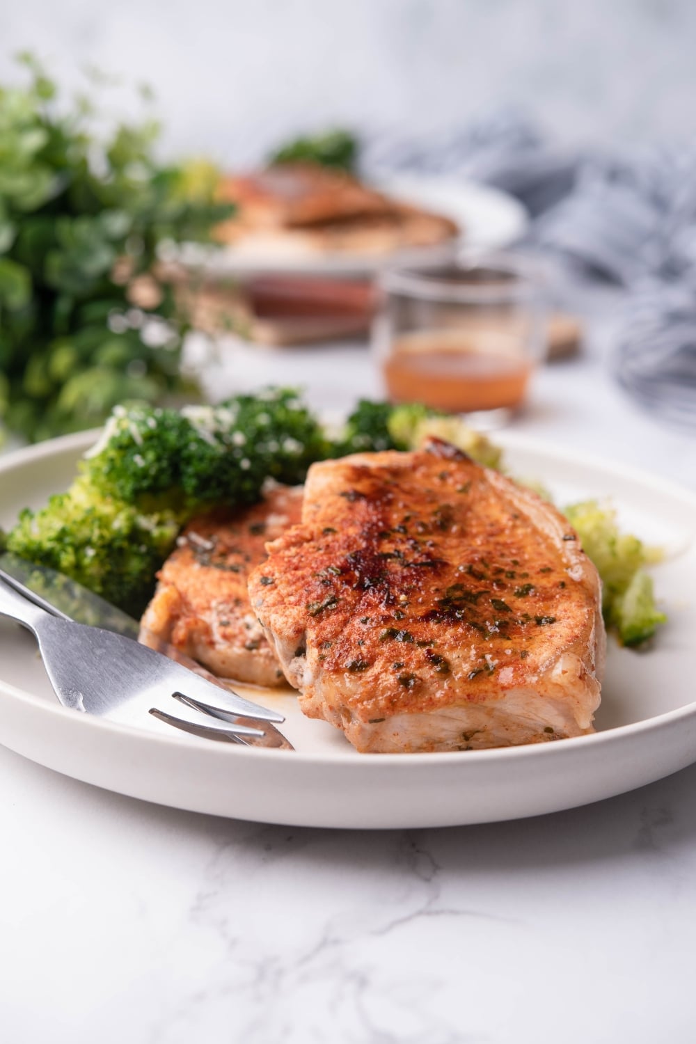 Sauteed pork chops served with steamed broccoli on a white plate, next to a mini pitcher of pan sauce. Another plate of sauteed pork chops with broccoli is in the back.