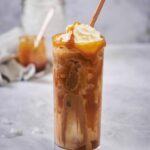 A tall glass of iced caramel macchiato with a brown straw. The coffee and milk has been mixed together and the glass is topped with whipped cream and caramel. Small pieces of melting ice are scattered on the countertop and jars of ice and caramel can be seen far in the back.