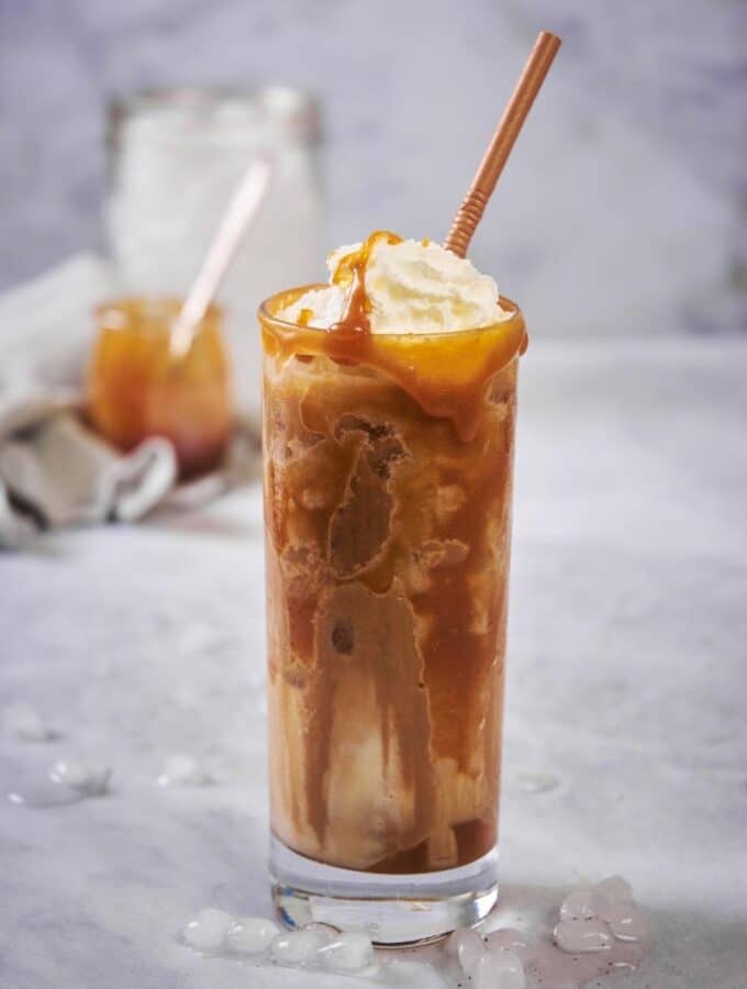 A tall glass of iced caramel macchiato with a brown straw. The coffee and milk has been mixed together and the glass is topped with whipped cream and caramel. Small pieces of melting ice are scattered on the countertop and jars of ice and caramel can be seen far in the back.