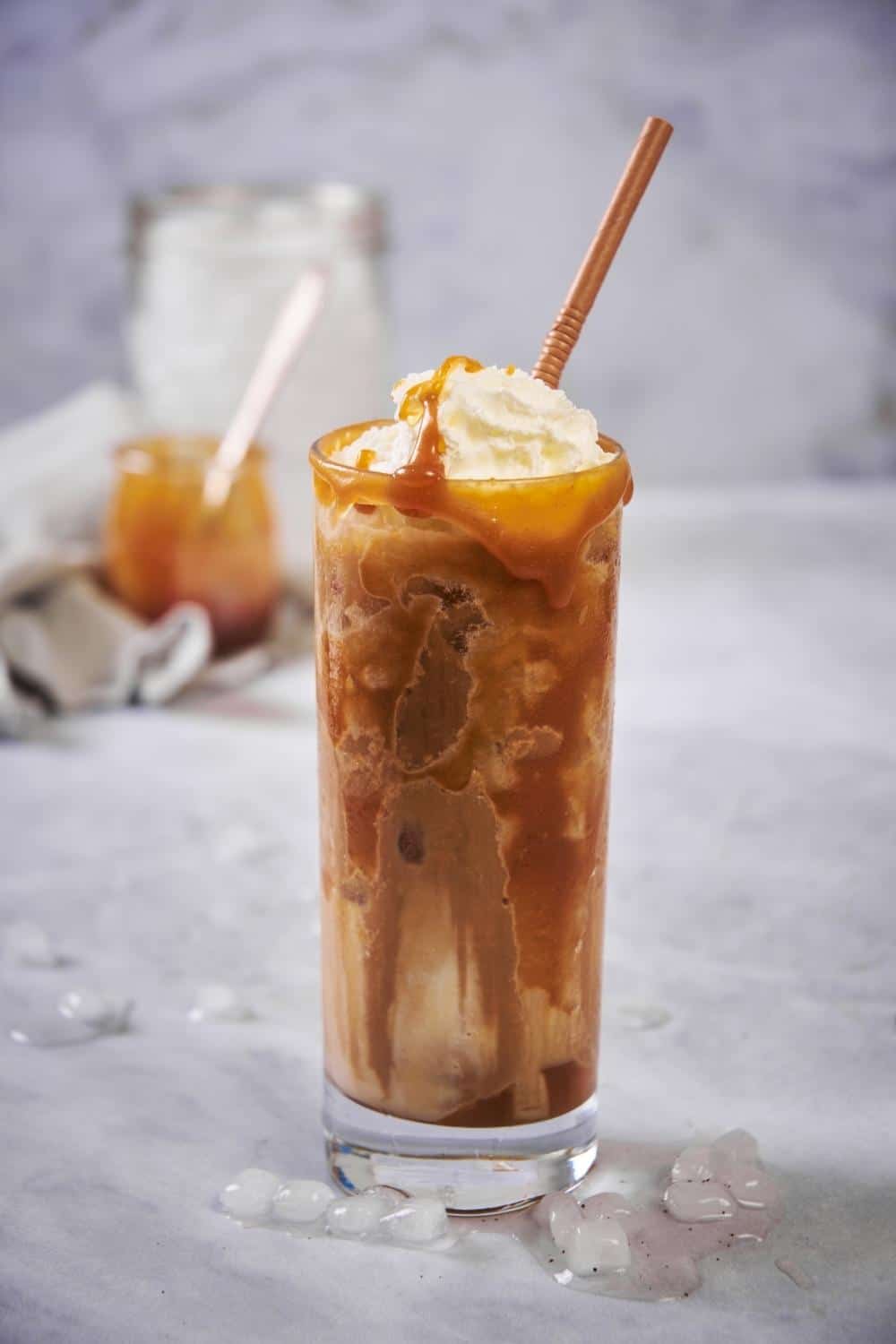 A tall glass of iced caramel macchiato with a brown straw. The coffee and milk has been mixed together and the glass is topped with whipped cream and caramel. Small pieces of melting ice are scattered on the countertop and jars of ice and caramel can be seen far in the back.
