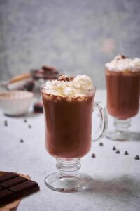 A glass mug of hot chocolate garnished with whipped cream and chocolate shavings. Another glass of hot chocolate with whipped cream can be seen in the back.