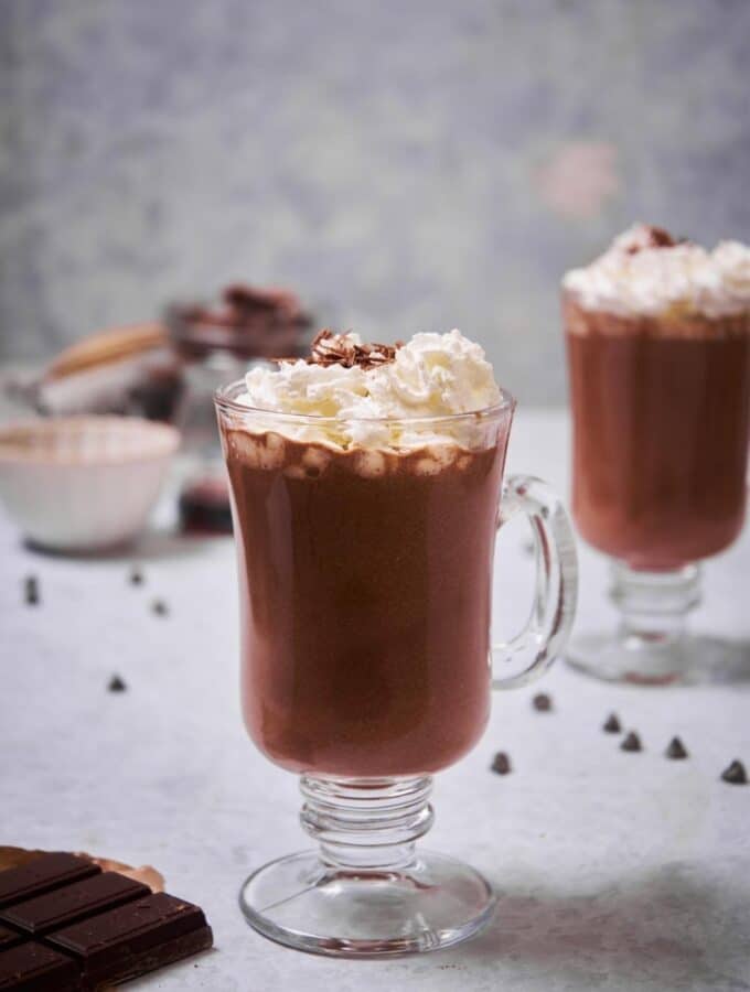 A glass mug of hot chocolate garnished with whipped cream and chocolate shavings. Another glass of hot chocolate with whipped cream can be seen in the back.
