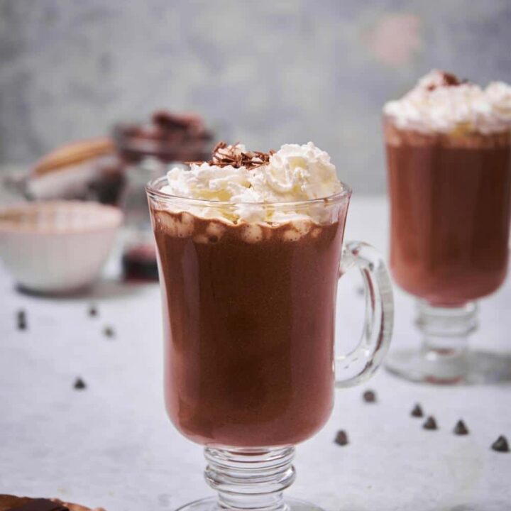 A glass mug of hot chocolate garnished with whipped cream and chocolate shavings. Another glass of hot chocolate with whipped cream can be seen in the back.