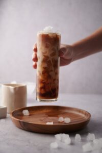 A hand picking up a tall glass of iced vanilla latte from a wooden tray scattered with bits of ice.