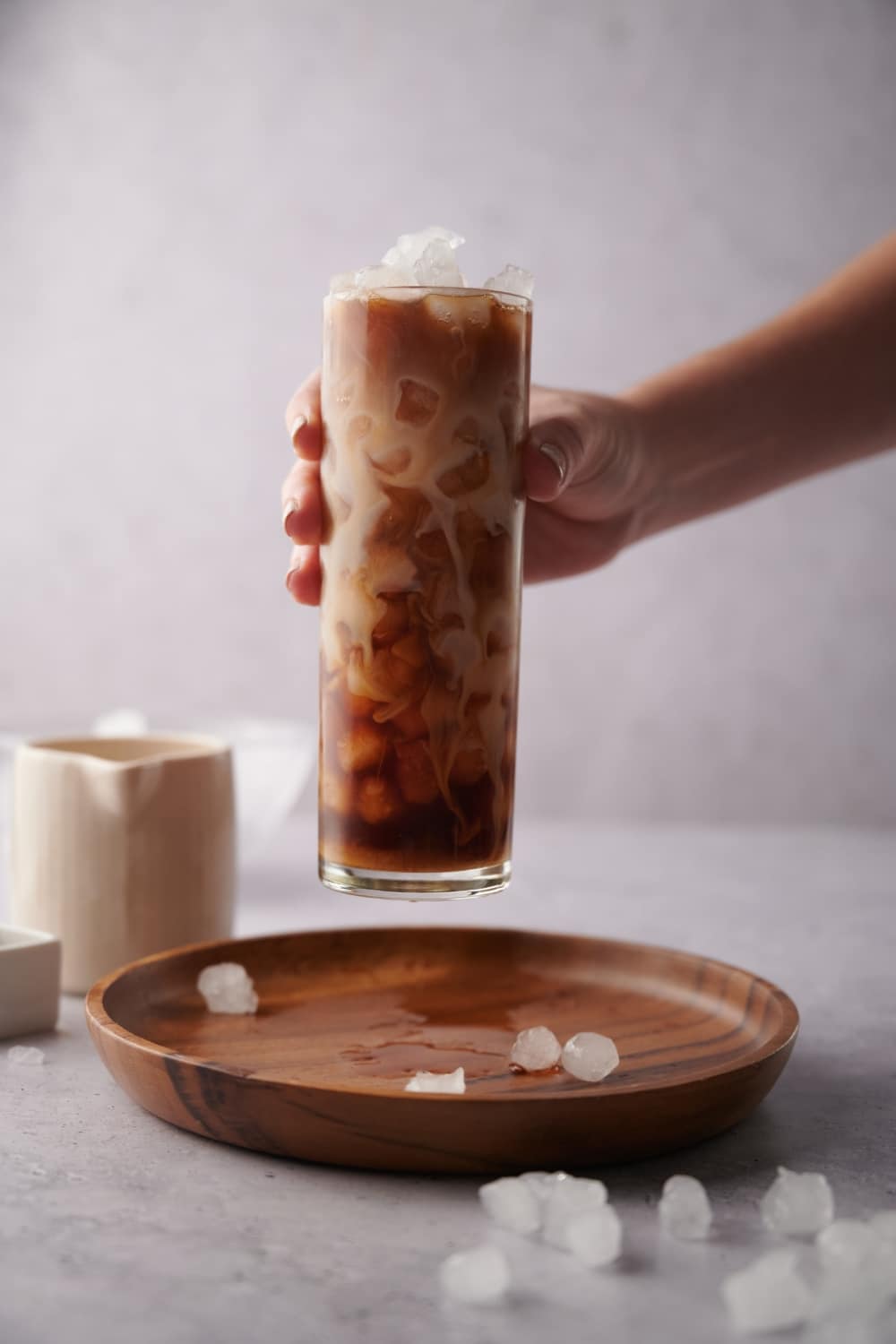 A hand picking up a tall glass of iced vanilla latte from a wooden tray scattered with bits of ice.