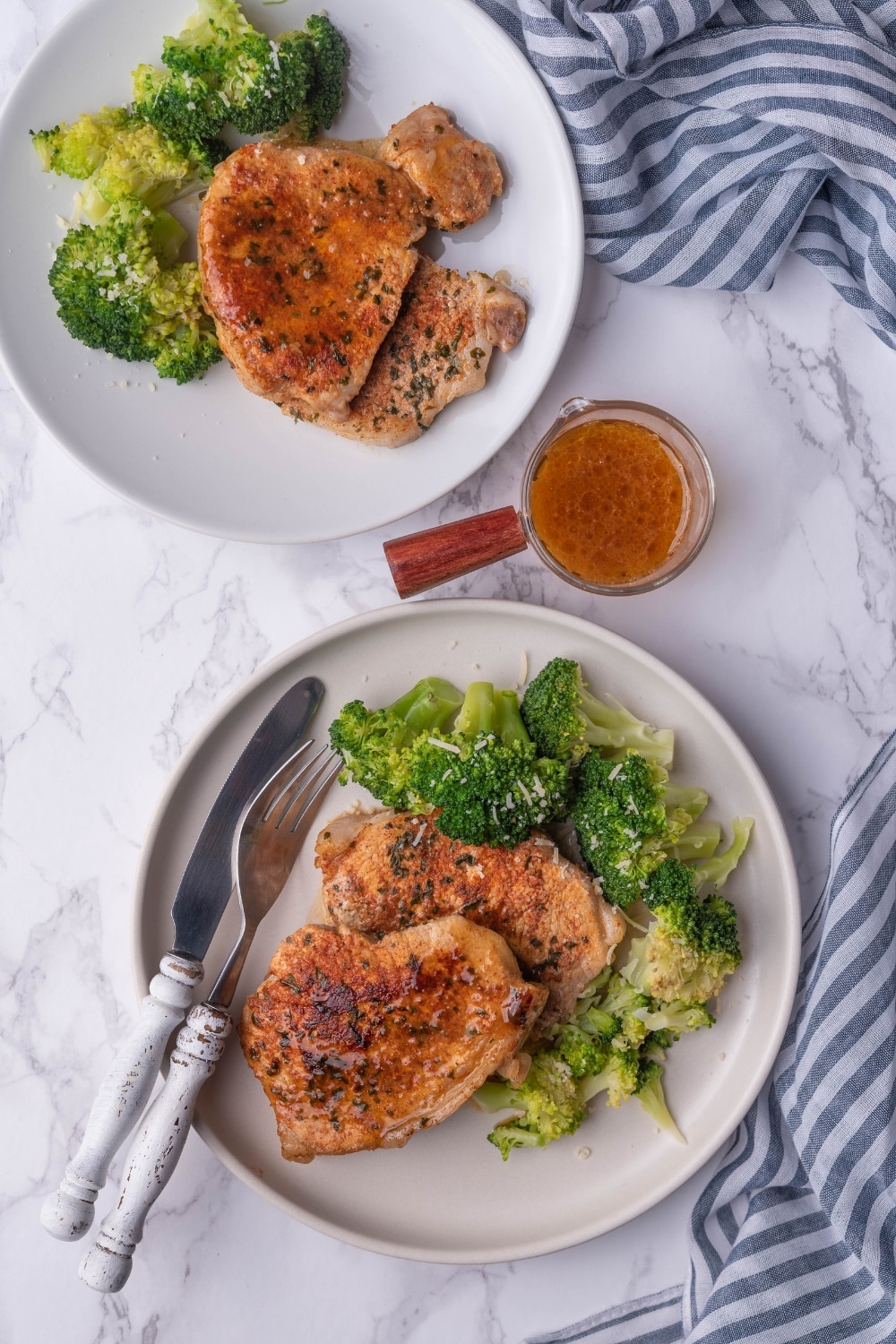 Two white plates each with two sauteed pork chops paired with steamed broccoli. Between the two plates is a mini pitcher of pan sauce.