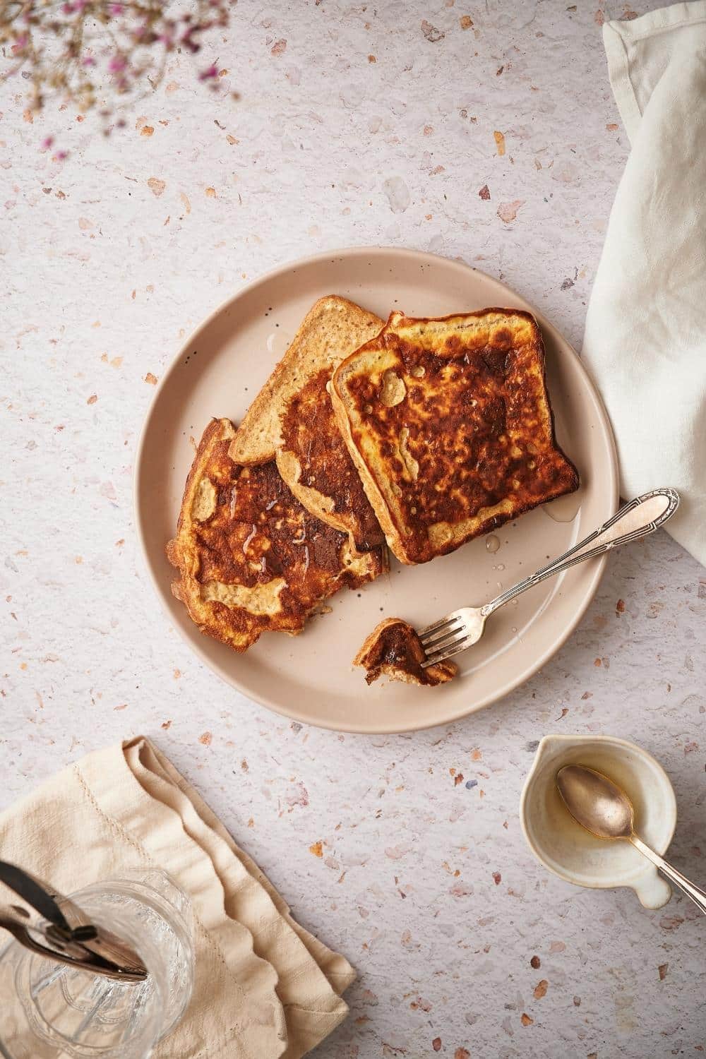 A plate of three french toasts fanned out on a plate. A metal fork with a piece of french toast sits on the plate and next to the plate is a small syrup pitcher with a spoon.