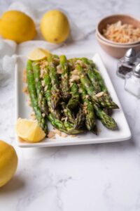 Sauteed asparagus garnished with panko breadcrumbs and served with lemon wedges on a rectangle white plate. Behind it are whole lemons on a tea towel, a small bowl of panko breadcrumbs, and salt and pepper shakers.