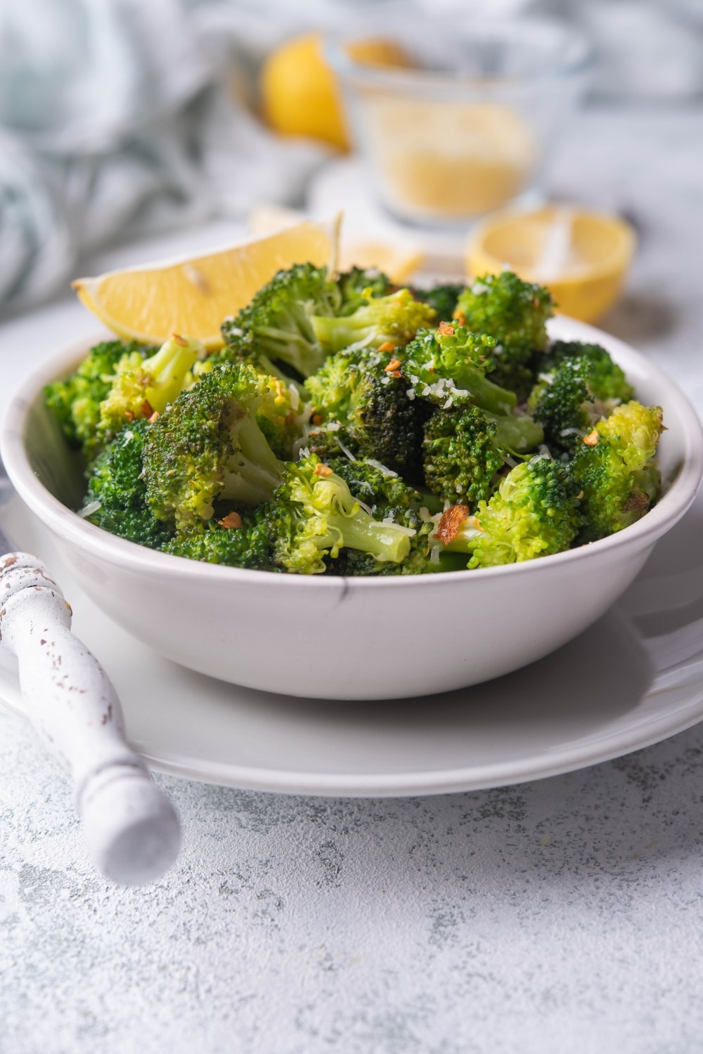 Sauteed broccoli with red pepper flakes and grated parmesan in a white bowl, served with a lemon wedge. The bowl sits on a white plate next to a fork with a white handle. In the back are more lemon slices.