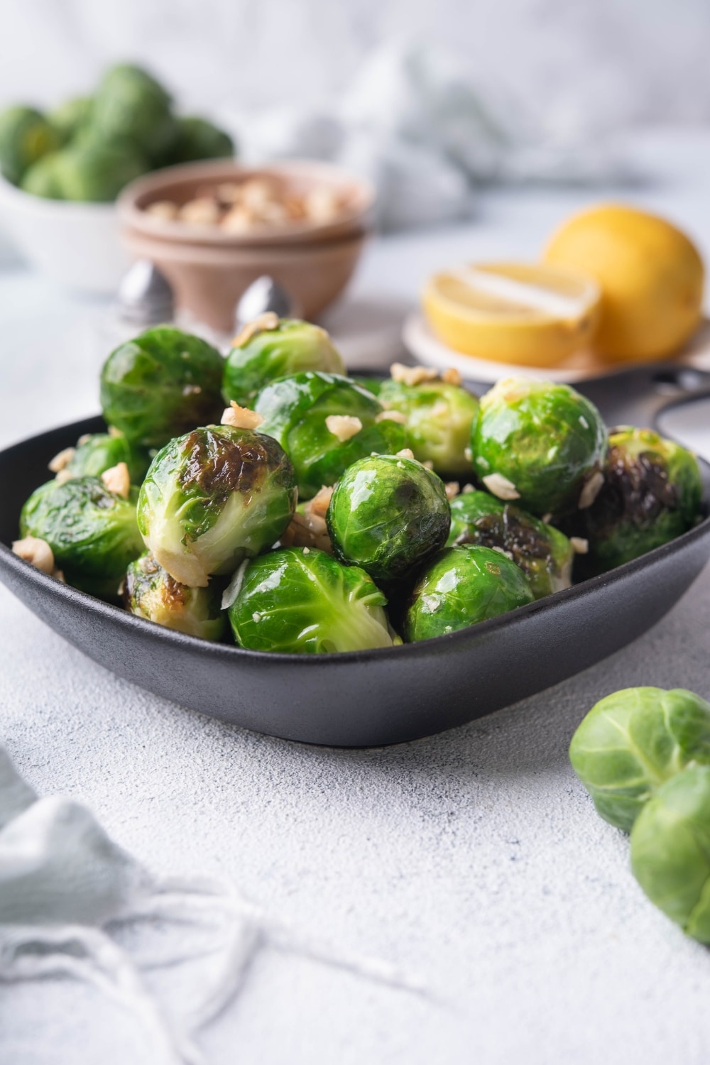 Sauteed brussels sprouts garnished with chopped hazlnuts in a small square cast iron skillet. Around the pan are whole uncooked brussel sprouts, a bowl of hazelnuts, salt and pepper shakers, and a plate of lemon with half a lemon wedge.