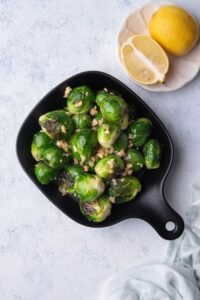 Top view of sauteed brussels sprouts with chopped hazelnuts served in a small square cast iron skillet. Next to the skillet is a small cream-colored plate with a halved lemon.