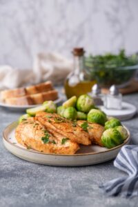 Sauteed chicken breasts garnished with herbs on a plate with steamed brussel sprouts. A folded striped tea towel is next to the plate. Behind it is a plate of sliced bread, a bottle of olive oil, salt and pepper shakers, and a glass bowl filled with greens.