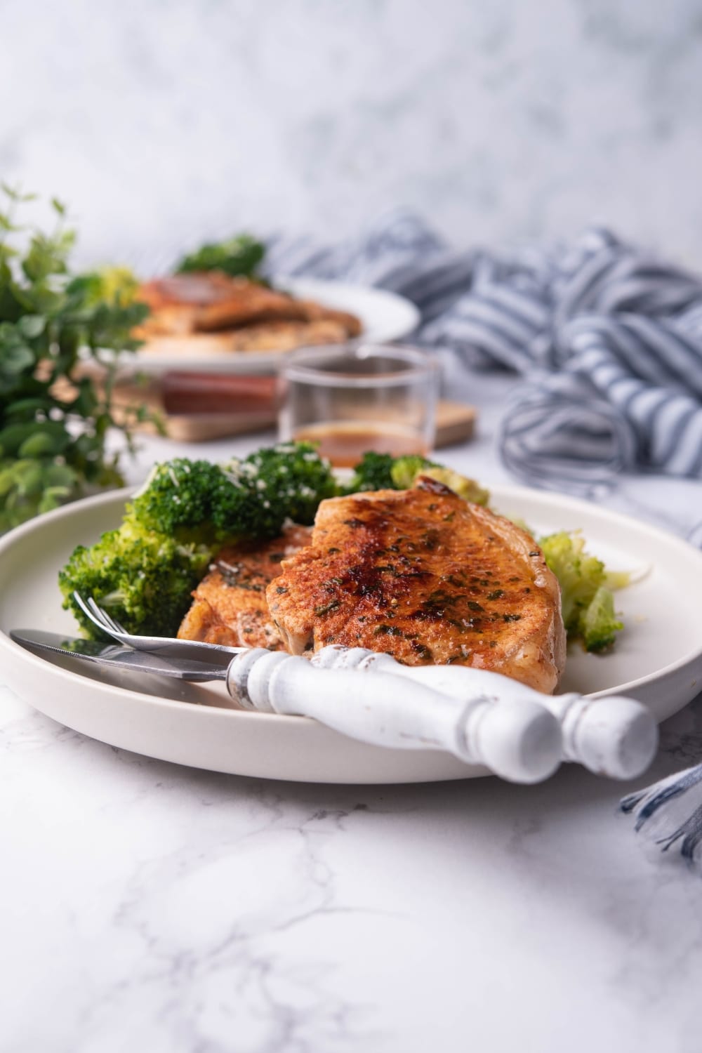 Thick pan seared pork chop with broccoli on a white plate with a fork and knife. Another plate of pan seared pork chops and broccoli can be seen in the background next to a mini glass pitcher of pan sauce.