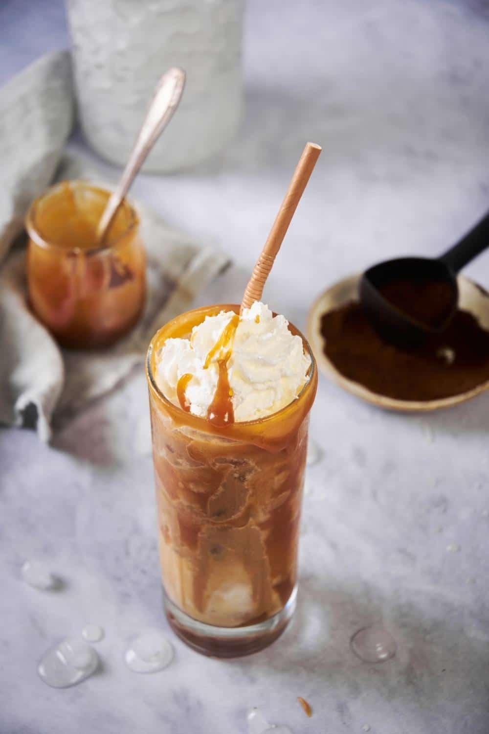 A tall glass of iced caramel macchiato topped with whipped cream and a brown straw. The coffee and milk are swirled together with caramel sauce drizzle lining the inside and rim of the glass. Behind it is a small plate with a spoonful of ground coffee, a jar of ice, and a small jar of caramel.