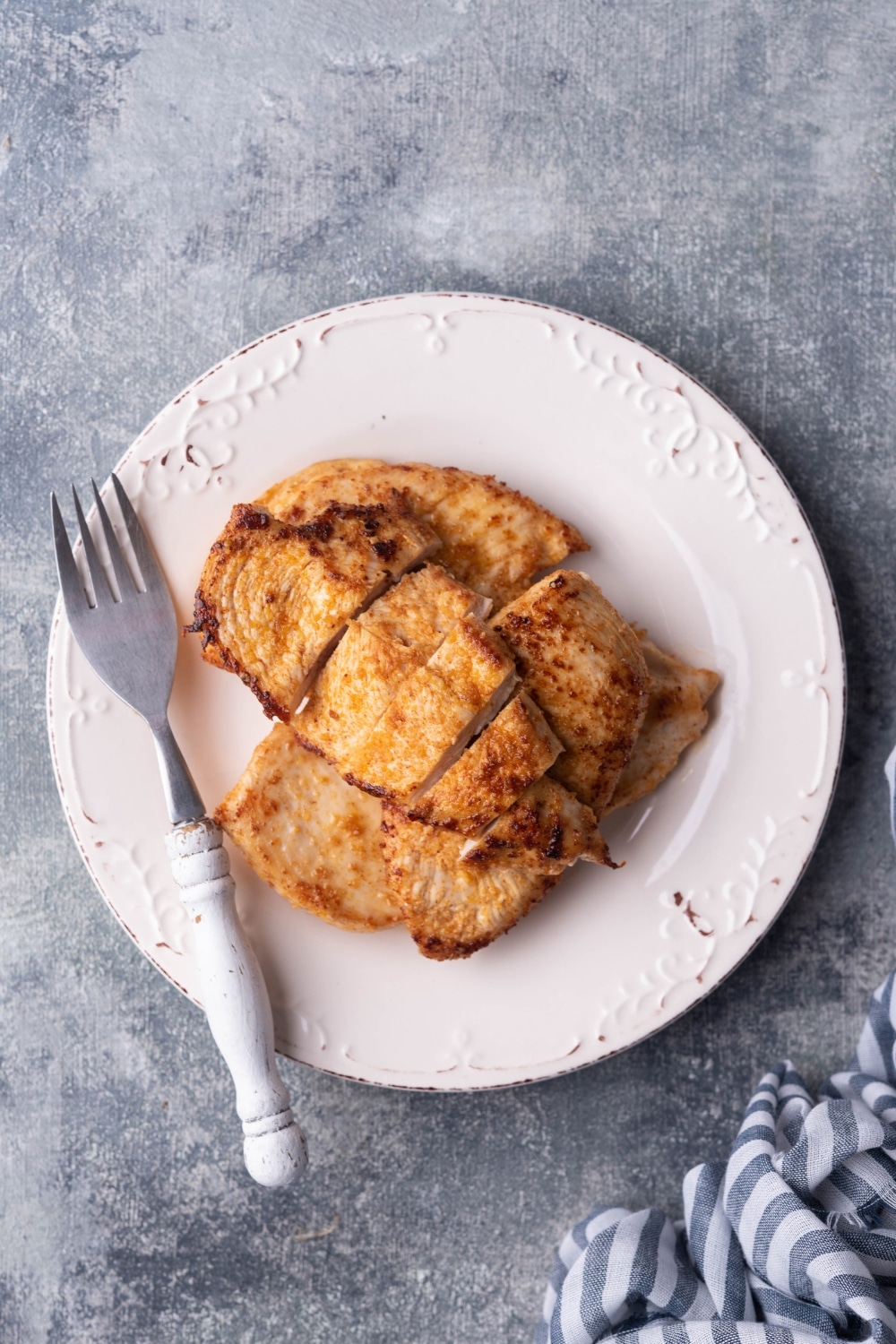 Sauteed chicken stacked on an embossed plate next to a fork with a white handle. The topmost chicken breast has been sliced into strips.