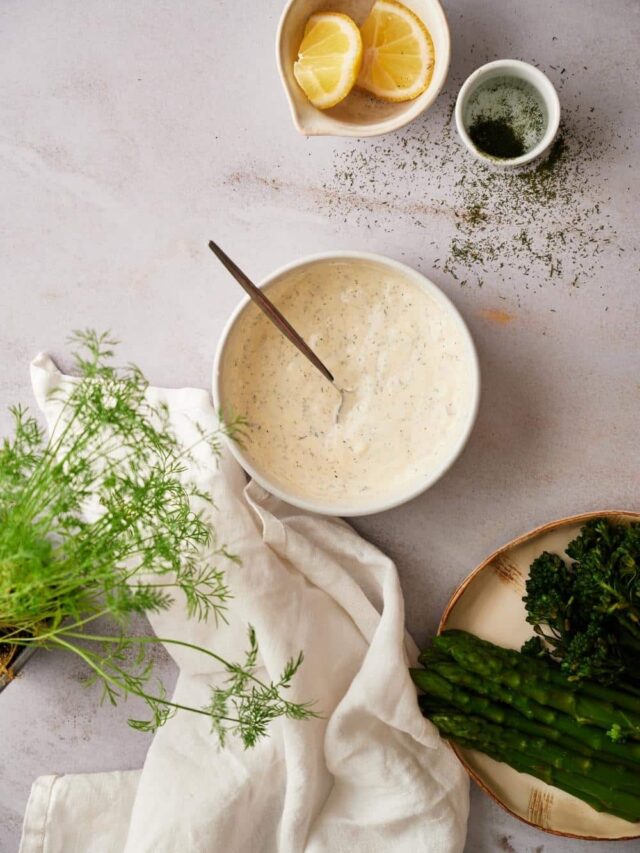 A medium sized bowl of dill sauce with a metal spoon resting in the center. Surrounding the bowl are small bowls of lemon wedges and chopped dill, a plate of asparagus with broccolini, a tea towel, and fresh dill fronds.