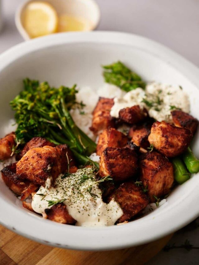 Air fryer salmon bites in a bowl of white rice, steamed broccolini, and steamed asparagus. The bowl is topped with two dollops of creamy dill sauce and garnished with fresh chopped dill. In the background is a small bowl of lemon wedges.