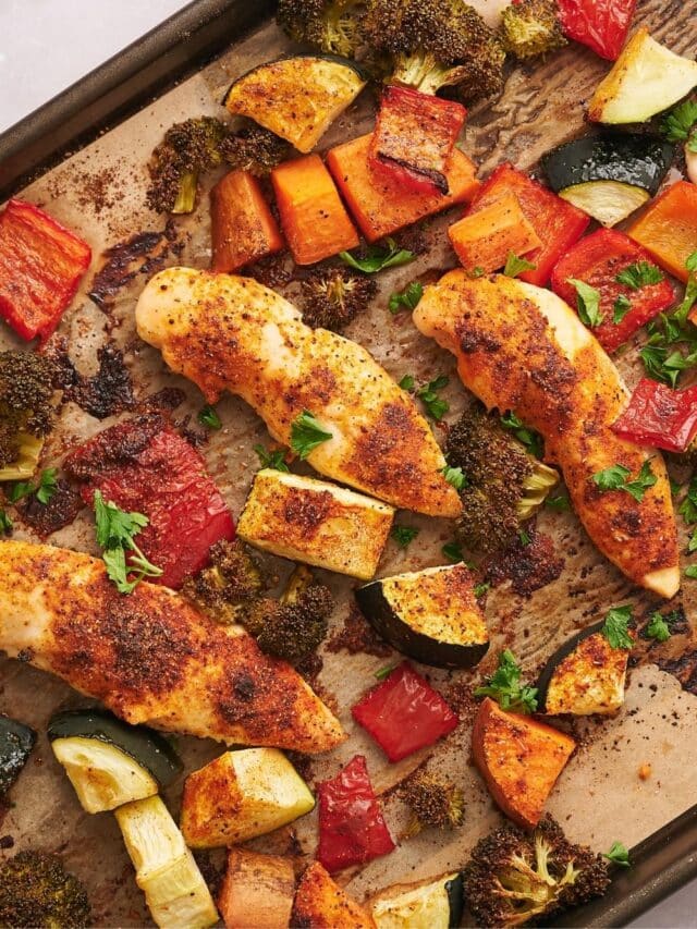 Three baked chicken tenderloins on a baking sheet lined with parchment paper surrounded by roasted broccoli, cucumber, and carrots.