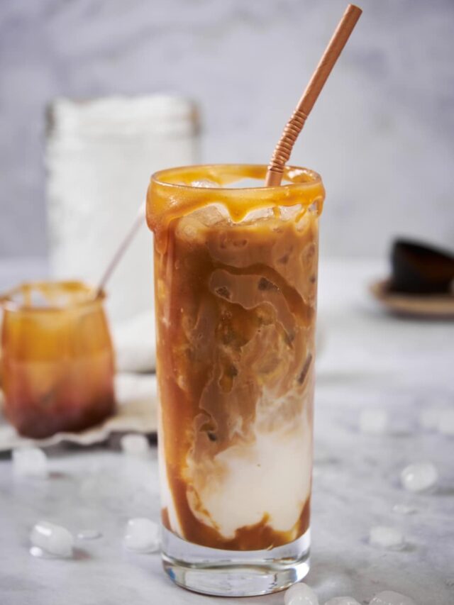 Keto iced caramel macchiato in a tall glass with a brown bendy straw. In the background is a small jar of of keto caramel with a spoon, a large mason jar of ice, and a measuring scoop of ground coffee on a small plate.