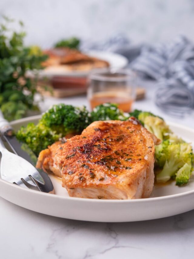 Pan seared pork chops with broccoli on a white plate. A white handled fork and knife are on the plate and another plate of pork chops and broccoli can be seen in the background next to a glass sauce pitcher of pan sauce.