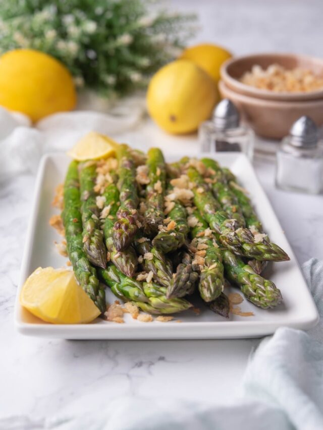 Close up of sauteed aspargus spears on a white rectangle plate. They're sprinkled with panko breadcrumbs and served with lemon wedges. In the back are whole lemons, salt and peppers shakers, and a bowl of breadcrumbs.