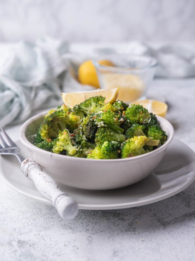 Sauteed broccoli florets with red pepper flakes and grated parmesan in a white bowl garnished with a lemon wedge. The bowl is on a white plate next to a fork with a white handle. Behind it are more lemon wedges and a gently crumpled tea towel.