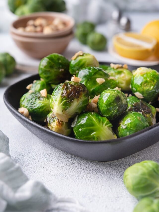 Close up of sauteed brussels sprouts in a small square cast iron skillet. The brussels sprouts are garnished with chopped hazelnuts. Behind them is a bowl of fresh raw brussel sprouts, a bowl of hazelnuts, salt and pepper shakers, and a plate of lemon with half a lemon wedge.