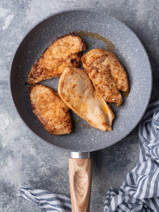 Four slices of sauteed chicken breasts in a grey speckled skillet with a wooden handle.