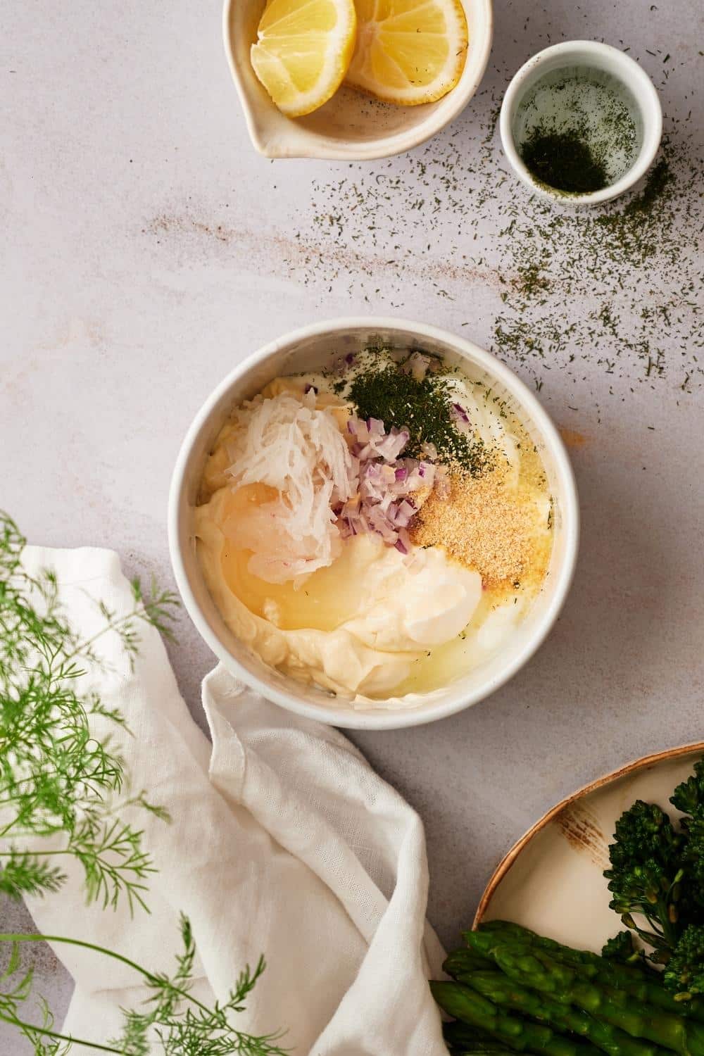 A bowl with unmixed dill sauce ingredients: yogurt, mayonnaise, garlic powder, horseradish, minced shallots, lemon juice, and chopped dill.