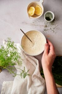 A hand holding a bowl of fresh dill sauce with a metal spoon. The bowl is surrounded by small bowls of lemon wedges and chopped dill, a plate of asparagus with broccolini, a tea towel, and fresh dill fronds.