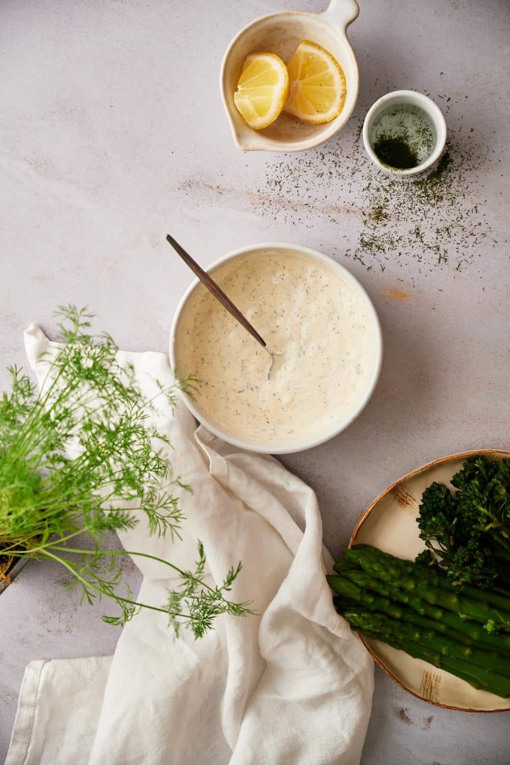A medium sized bowl of dill sauce with a metal spoon resting in the center. Surrounding the bowl are small bowls of lemon wedges and chopped dill, a plate of asparagus with broccolini, a tea towel, and fresh dill fronds.