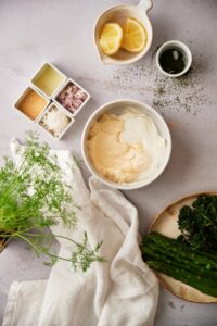 A medium sized bowl filled with yogurt and mayonnaise. The bowl is surrounded by prepared dill sauce ingredients and part of a plate of broccolini and asparagus can be seen on the side.