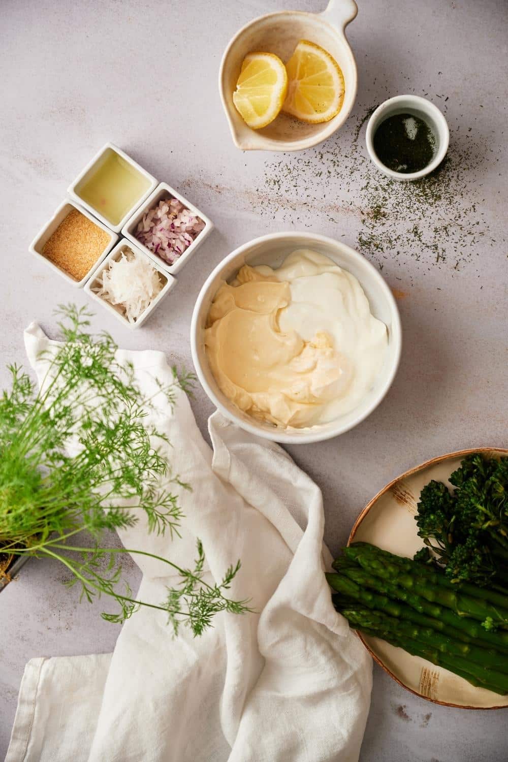 A medium sized bowl filled with yogurt and mayonnaise. The bowl is surrounded by prepared dill sauce ingredients and part of a plate of broccolini and asparagus can be seen on the side.