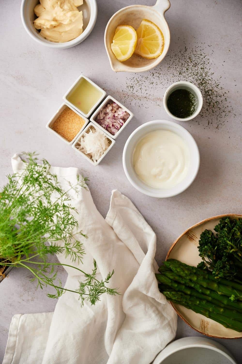 Dill sauce ingredients: small bowls of mayonnaise, yogurt, and lemon wedges; tiny bowls of chopped dill, minced shallots, garlic powder, lemon juice, and horseradish. At the bottom is a plate of asparagus and broccolini.