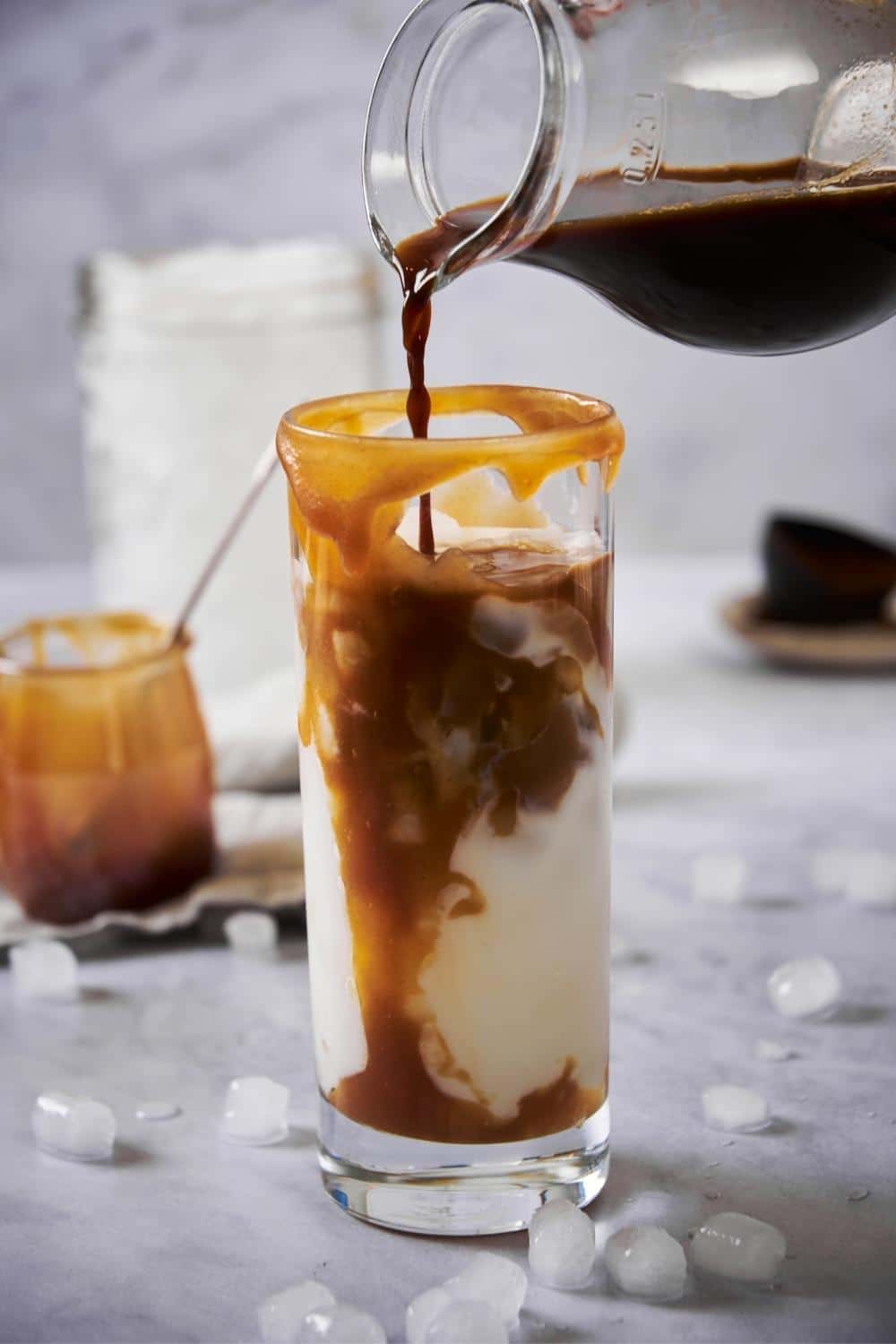 Cold brew coffee being poured from a glass pitcher into a tall glass of milk decorated with caramel syrup. In the back is a large jar of ice and a small jar of caramel, and small pieces of ice are scattered around the counter.