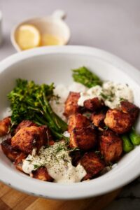 Air fryer salmon bites in a bowl of white rice, steamed broccolini, and steamed asparagus. The bowl is topped with two dollops of creamy dill sauce and garnished with fresh chopped dill. In the background is a small bowl of lemon wedges.