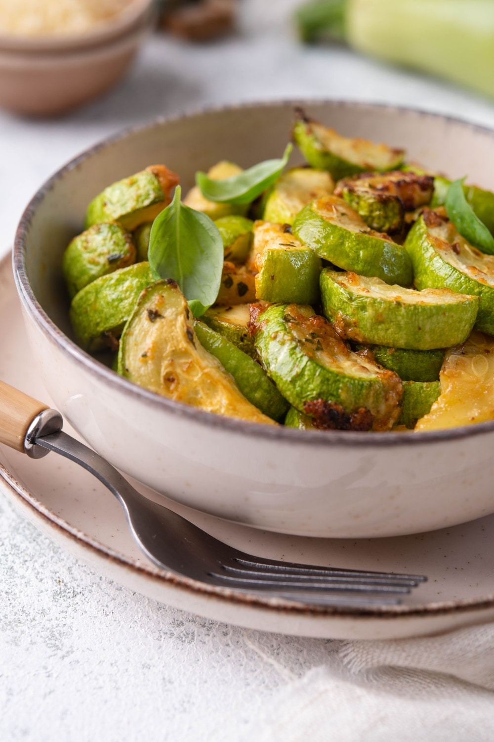 Close up of air fried zucchini garnished with fresh herbs in a bowl. The bowl is on plate with a wooden handle fork. Surrounding the plate is a fresh zucchini, a small bowl of grated parmesan, and a tea towel.