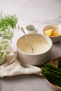 A medium white bowl of freshly mixed dill sauce. Behind it is a small bowl of lemon wedges and a tiny bowl of dill. In front of it is a plate of asparagus and broccolini and a tea towel with fresh dill fronds.