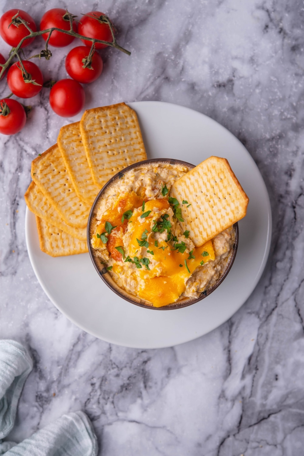 Top view of a bowl of chicken dip with a cracker dipped in it. The bowl is on a plate with more crackers and next to it are cherry tomatoes on the stem.