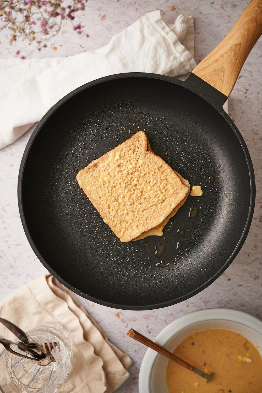 A slice of french toast cooking in a black pan with a wooden handle. Next to the pan is a bowl of french toast batter.
