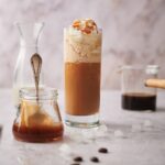 Keto frappuccino in a tall skinny glass topped with whipped cream and caramel sauce. Surrounding it is a jar of caramel sauce with a spoon, a half filled carafe of almond milk, and a glass coffee pitcher half filled with coffee.