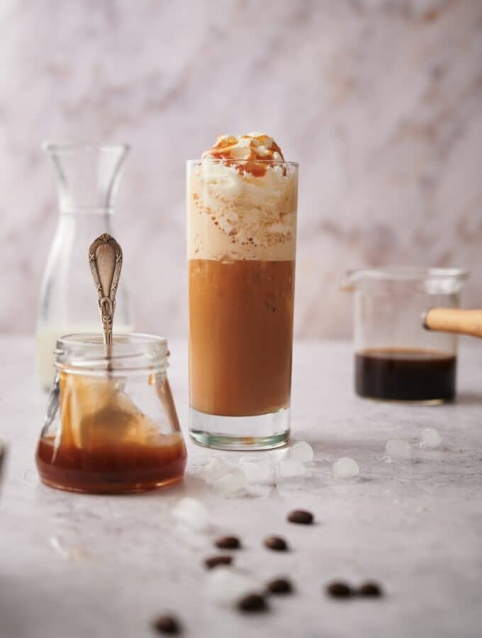 Keto frappuccino in a tall skinny glass topped with whipped cream and caramel sauce. Surrounding it is a jar of caramel sauce with a spoon, a half filled carafe of almond milk, and a glass coffee pitcher half filled with coffee.