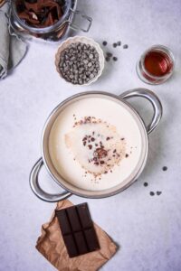 A saucepan filled with milk. Melting chocolate chips sit in the center of the milk. Surrounding the saucepan is a jar of chopped chocolate, a small bowl of chocolate chips, a small glass container of maple syrup, and a piece of baking chocolate.