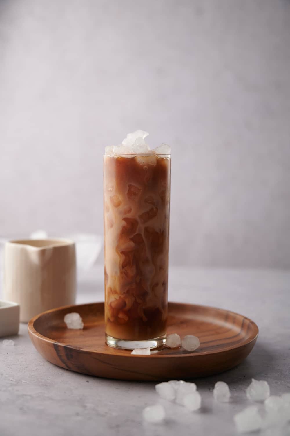 A tall glass of iced vanilla latte on a wooden tray. Small pieces of ice are scattered around the tray and a small ceramic milk pot is in the back.